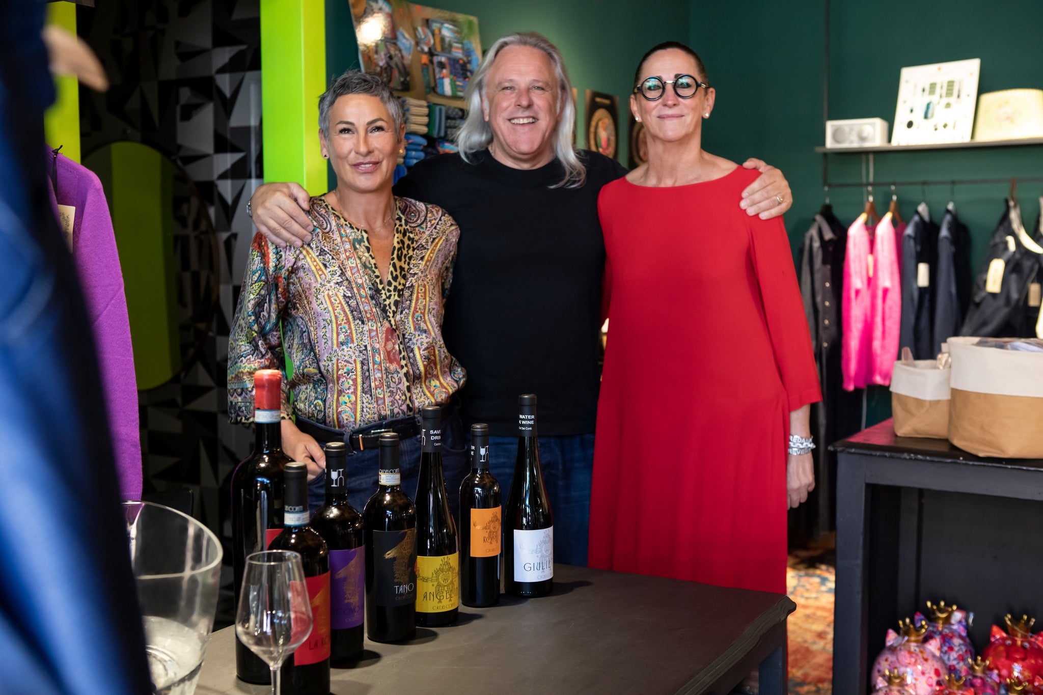 Three people posing together in a store with wine bottles and glasses on a table.