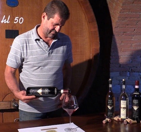 Man holding a bottle of wine in front of a wooden barrel with more bottles on a table.
